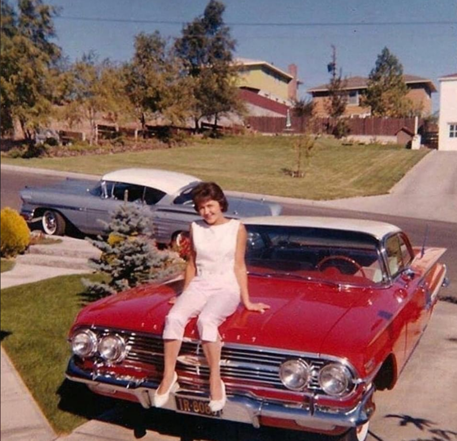 A woman in a white outfit sits smiling on the hood of a shiny red vintage car parked in a suburban driveway, with another classic car and houses visible in the background on a sunny day.