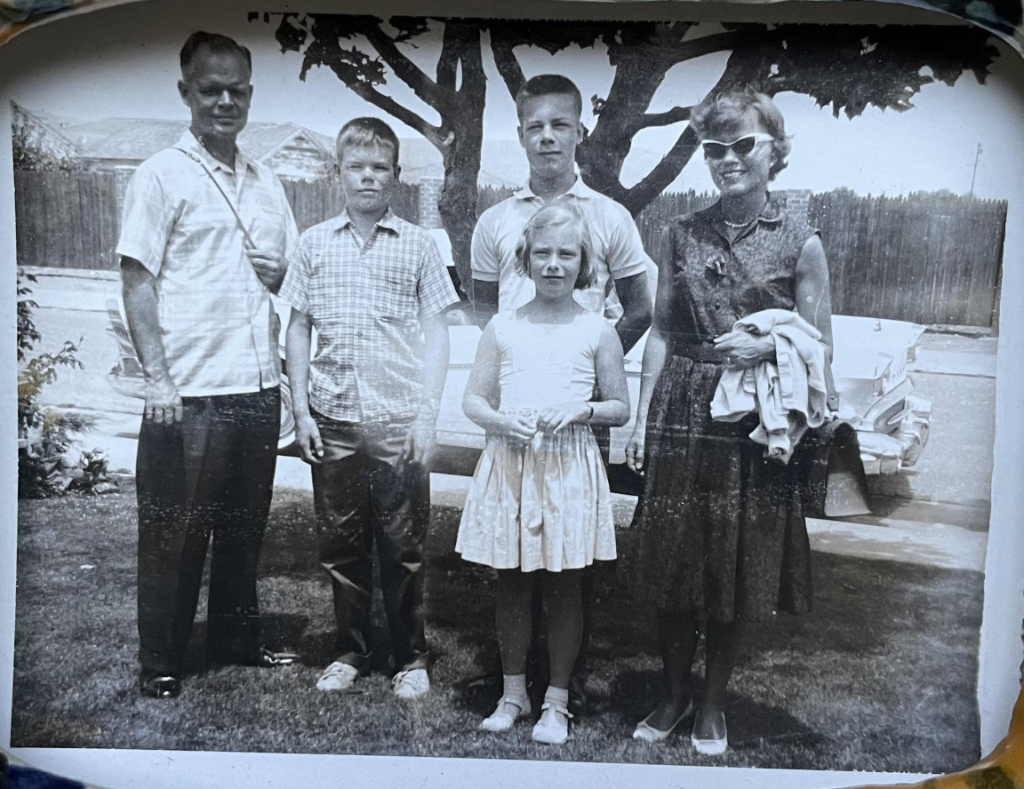 A vintage black-and-white photo of five people—two adult men, one adult woman, and two children—posing outdoors in front of a car and large tree. The woman wears sunglasses and holds a coat.