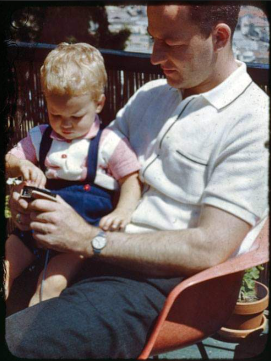 A man sits in a chair outdoors with a young child on his lap. The child, wearing suspenders and a short-sleeve shirt, focuses on an object in his hands while the man looks on attentively.