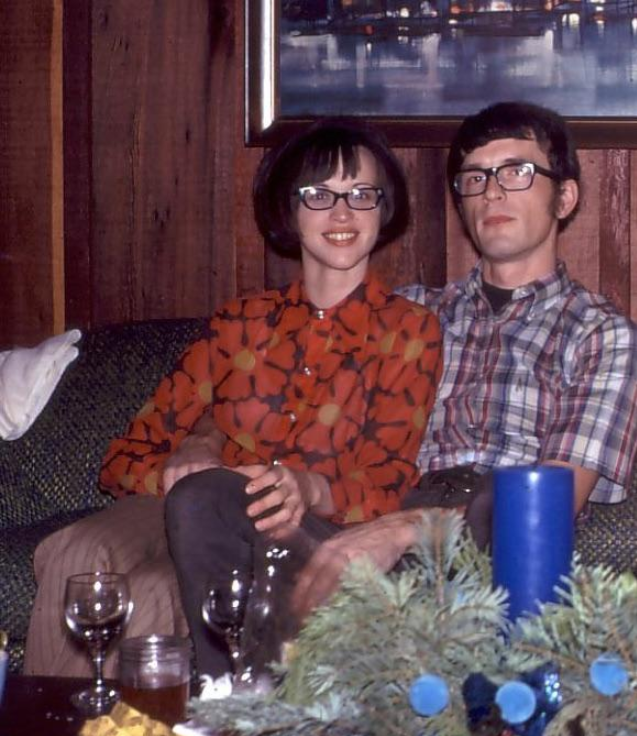 A woman and a man sit closely together on a patterned couch, both wearing glasses. She wears a red patterned blouse; he wears a plaid shirt. Wine glasses and a blue candle are on the table in front of them.