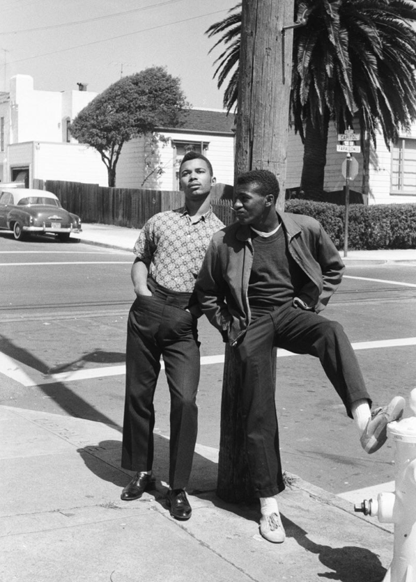 Two stylish young men stand on a street corner in front of a utility pole; one leans against the pole with a raised leg, palm trees, a vintage car, and houses are visible in the background.