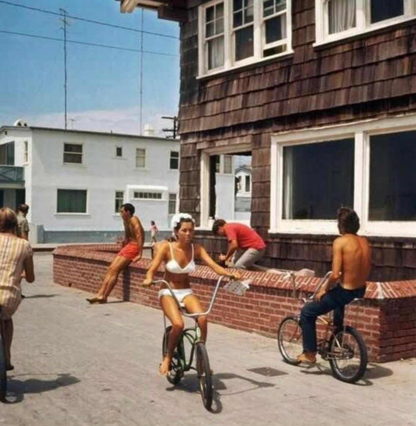 A woman in a white bikini rides a bicycle near a brown shingled building, while four men in swimsuits and shorts sit or stand nearby on a sunny day. Another man rides a bike shirtless.
