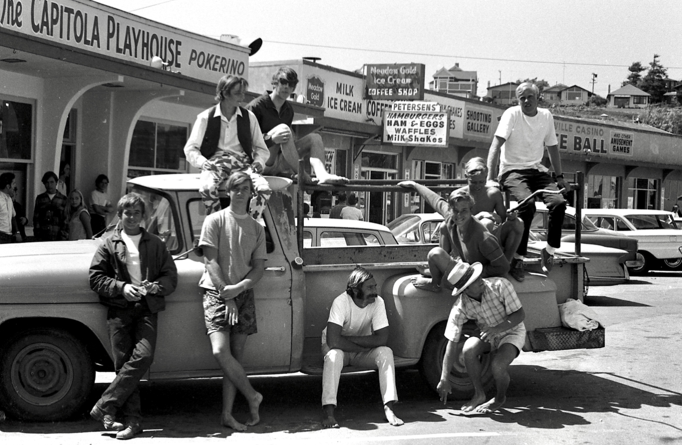 A group of people, mostly men, sit and stand on and around a pickup truck parked in front of shops in a busy street, some in casual summer clothing, with storefronts and cars visible in the background.