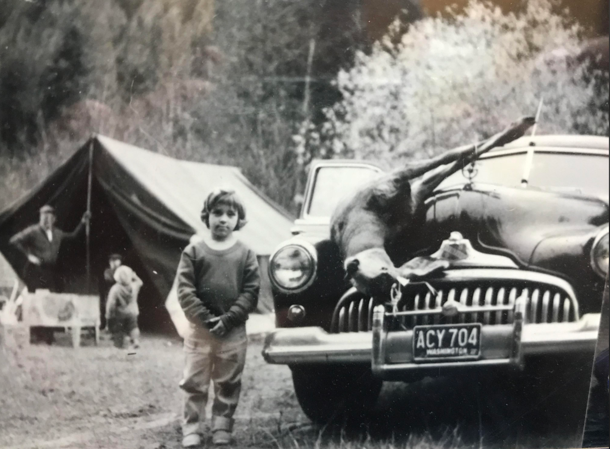 A black and white photo of a young child standing in front of a vintage car with a deer strapped to its hood. Behind them, a tent is set up outdoors, and a person stands near the tent.