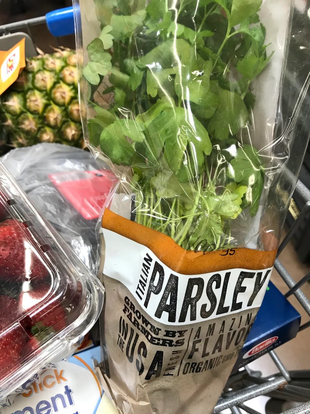 A package of Italian parsley, part of Walmart products, sits in a grocery cart alongside strawberries, a pineapple, and other assorted groceries. The parsley is in a clear plastic bag with brown and white labeling.