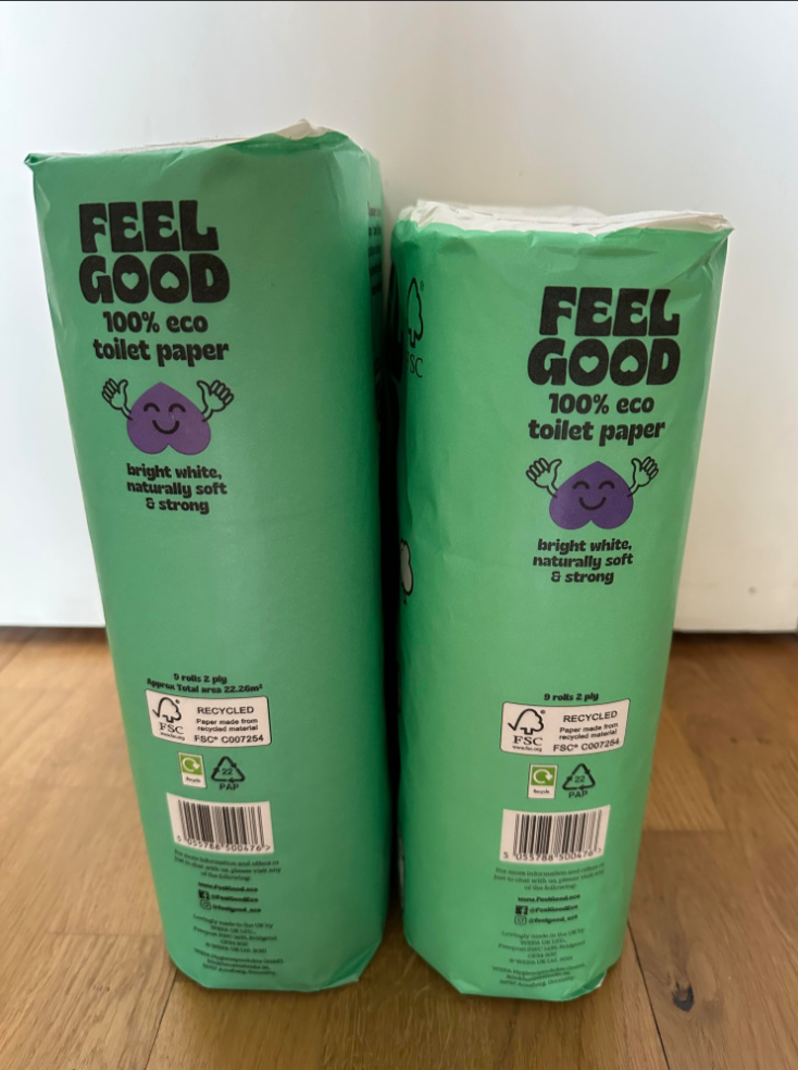 Two green packages of "Feel Good" 100% eco toilet paper stand upright on a wooden floor. The packaging highlights that the product is bright white, naturally soft, strong, and made from recycled materials.