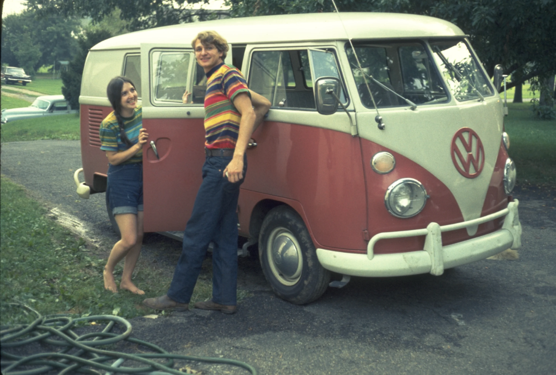 A young man and woman in striped shirts stand by an open door of a vintage red and white Volkswagen van, smiling outdoors on a driveway with green trees and grass nearby.