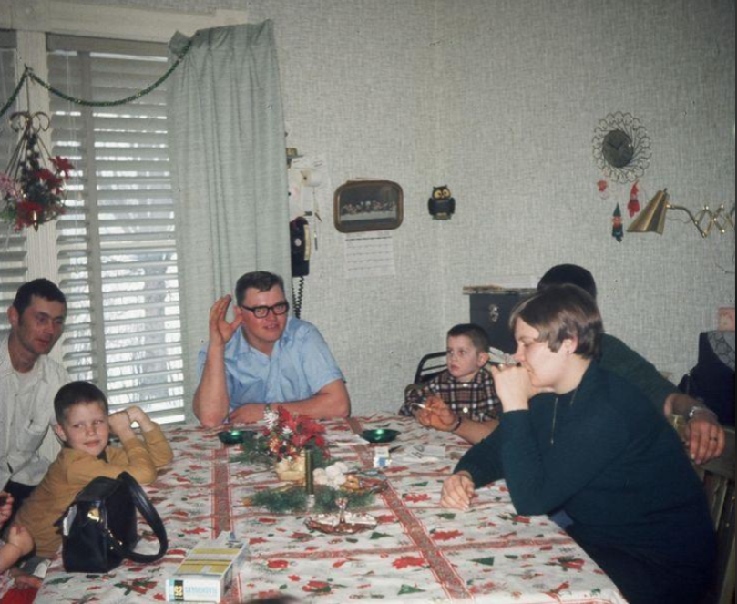 A family sits around a festive dining table with a holiday tablecloth, some eating and some talking. Four adults and three children are visible, with holiday decorations and a rotary phone on the wall in the background.