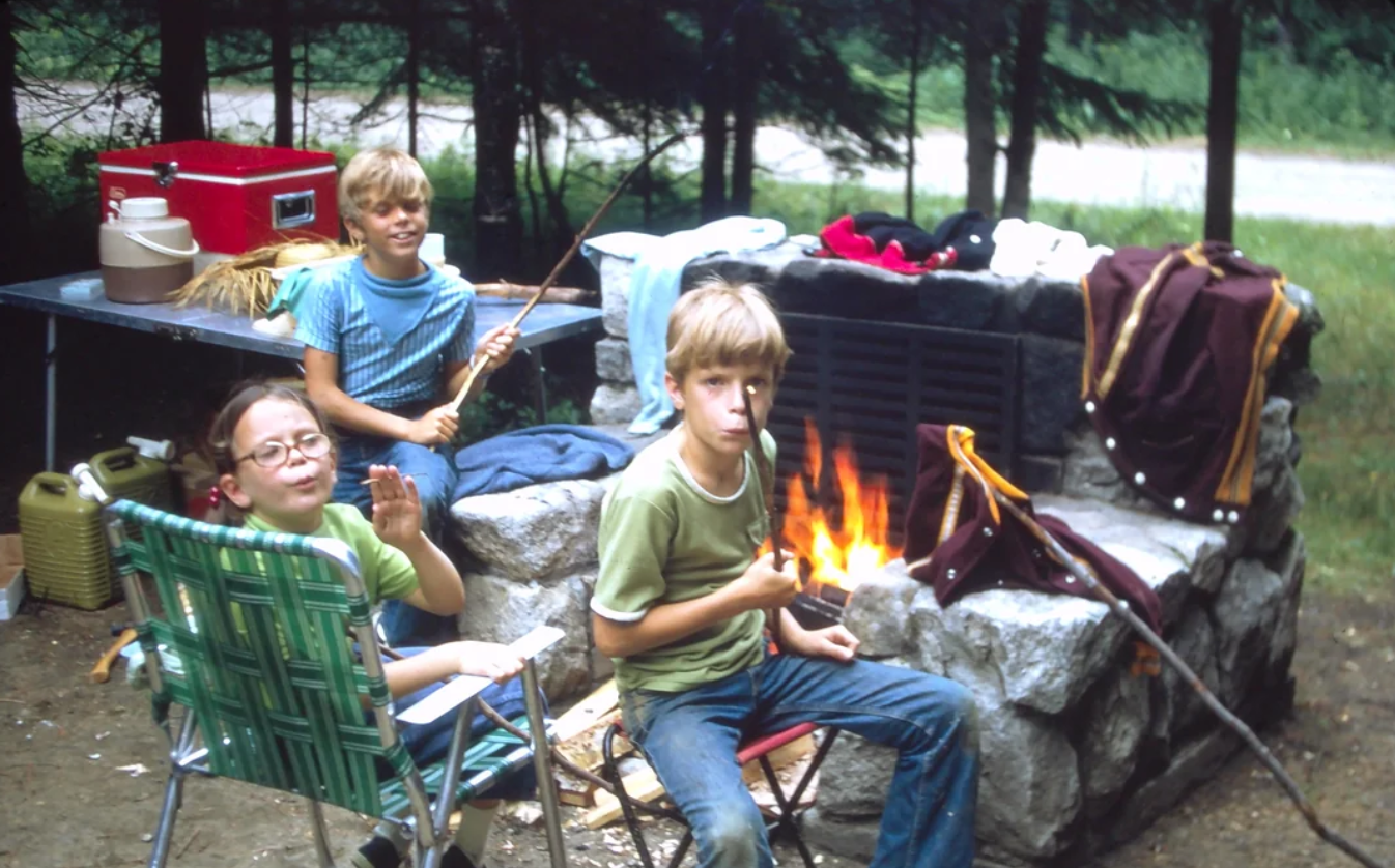 Three children sit around a campfire roasting food on sticks, with camping gear and a picnic table in the background, surrounded by trees and nature. One child waves and another looks at the camera.