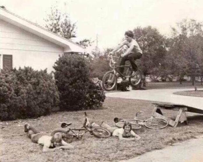 A person on a bicycle jumps off a wooden ramp over two people lying on the grass, with two other bikes nearby, outside a house surrounded by trees and bushes.