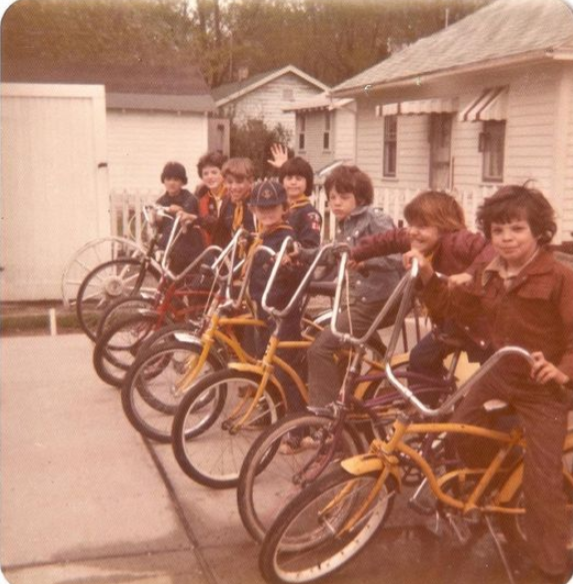 A group of eight children pose in a row on colorful bicycles in a driveway. They are smiling and some wave at the camera. Small white houses and green trees are visible in the background.