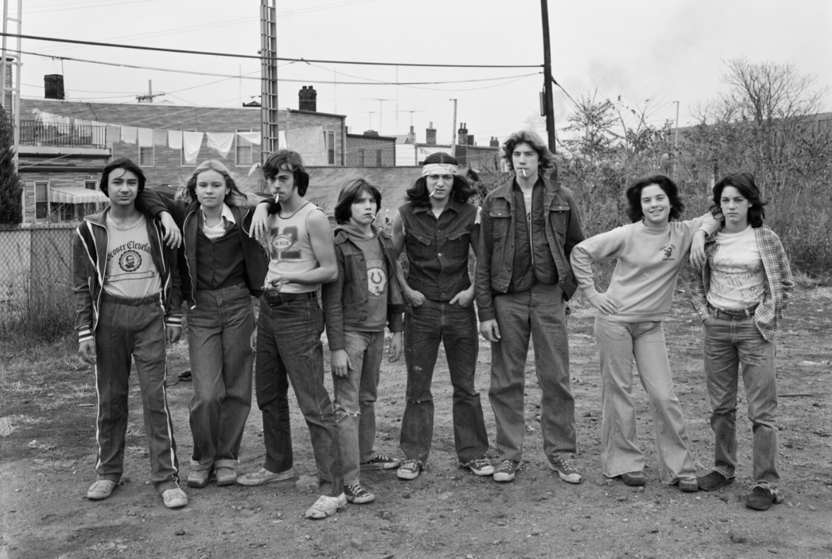 A group of eight teenagers in casual 1970s clothing stand together outside on a dirt lot, with houses, leafless trees, and utility poles in the background.