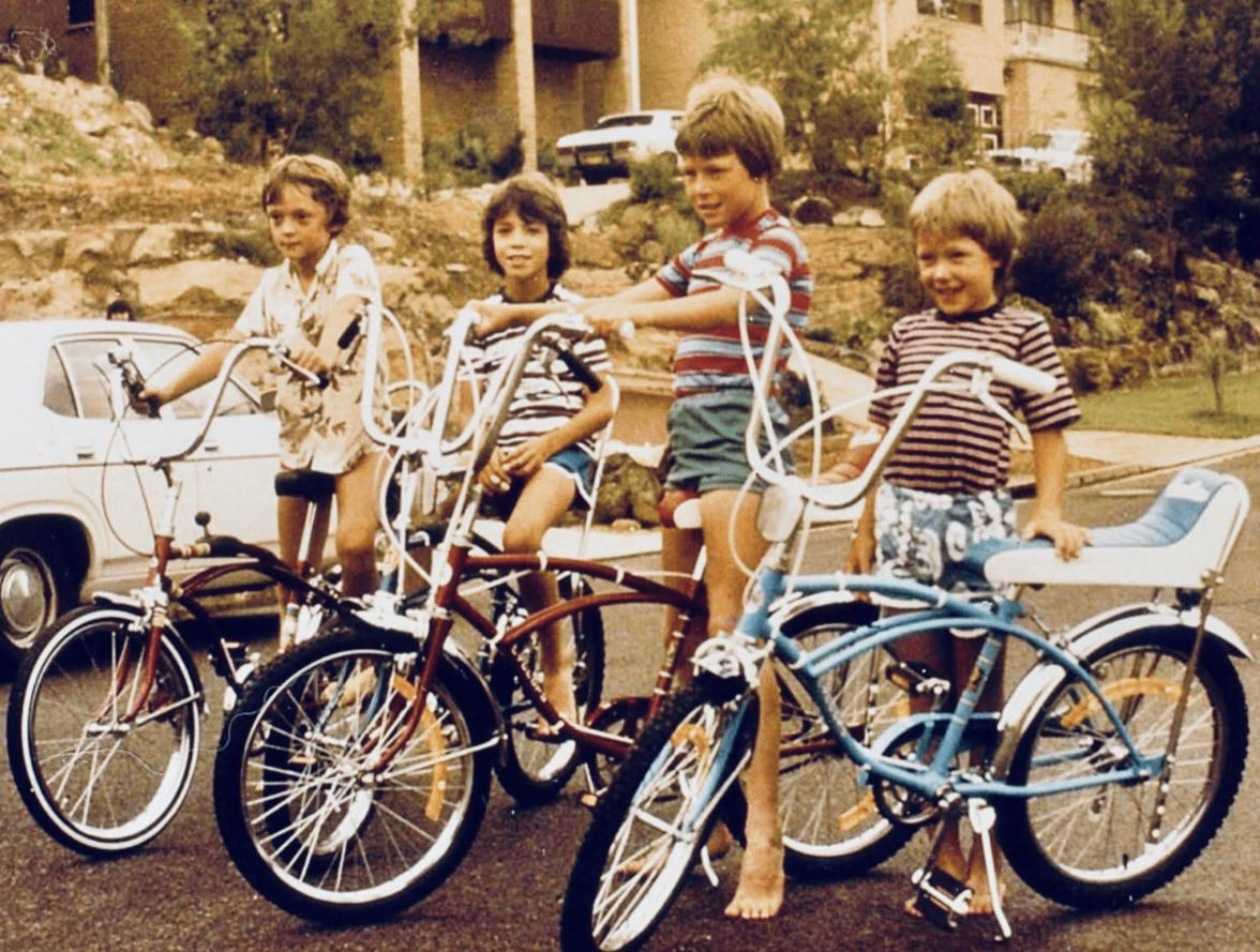 Four children stand outdoors on a street with vintage bicycles, smiling. They wear casual summer clothes, and there is a white car and houses in the background. The scene appears to be from the 1970s or 1980s.