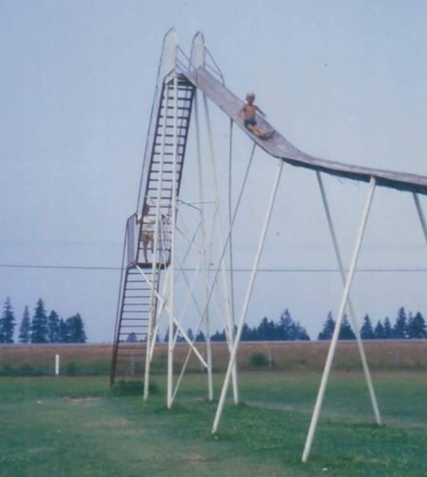 Two children play on an extremely tall and steep metal slide in a grassy field; one is climbing the ladder while the other sits at the top, with trees and a blue sky in the background.