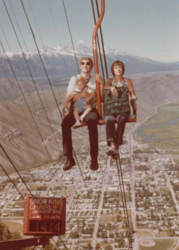 A man, woman, and young child ride a chairlift high above a town with mountains in the background. A sign below reads "Snow King Chairlift, Jackson, Wyo, June 30, 1974, 11:14.