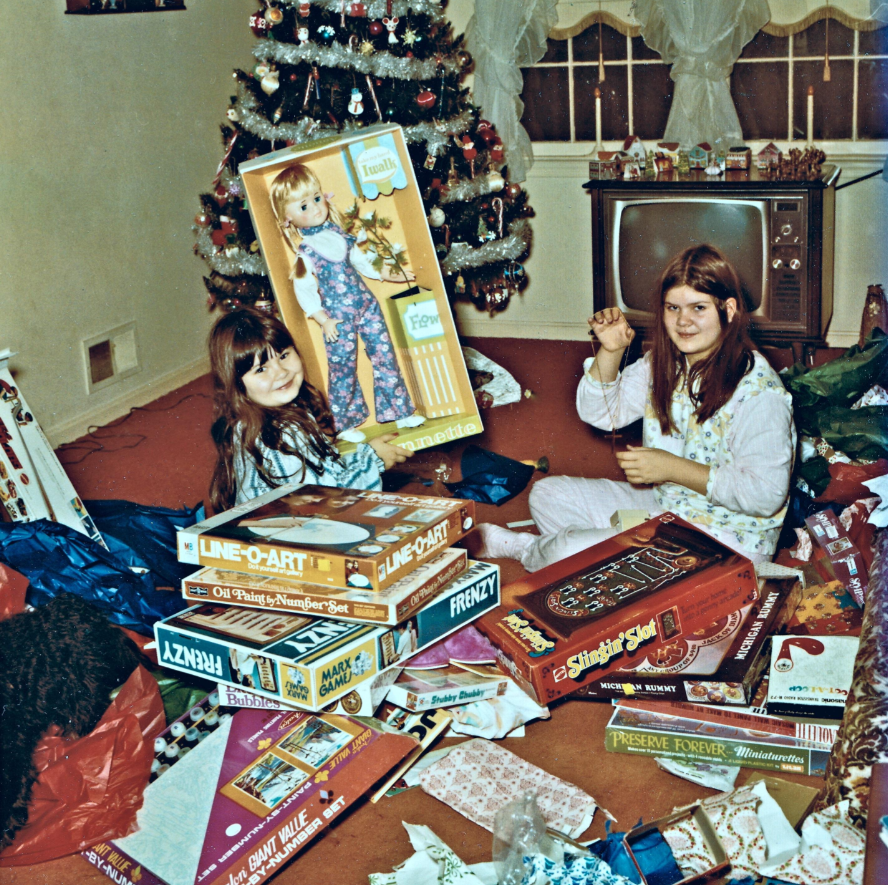 Two young girls sit on a carpet surrounded by opened presents and board games in front of a decorated Christmas tree and vintage TV, smiling and enjoying Christmas morning.