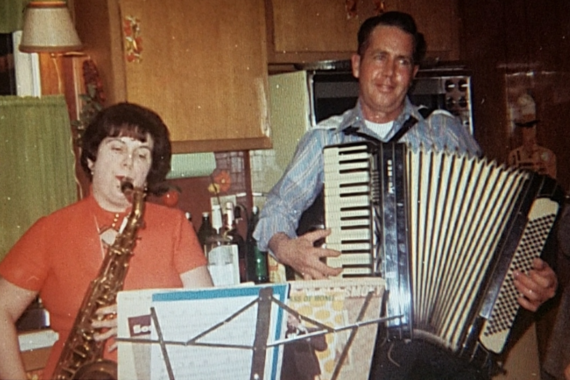 A woman plays a saxophone and a man plays an accordion in a kitchen. Both are smiling, with music stands holding sheet music in front of them. Bottles and kitchen items are visible in the background.