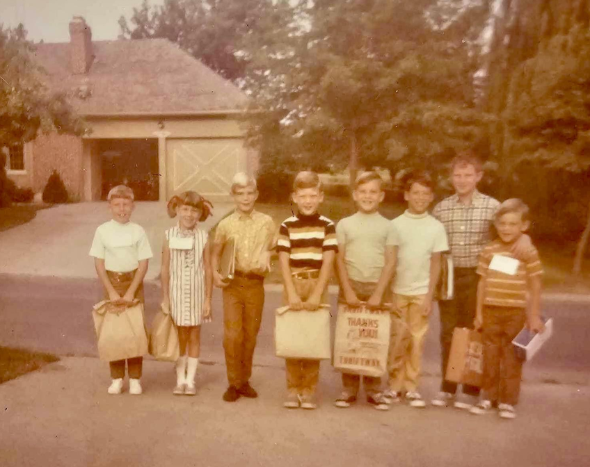 Eight children stand in a row outside on a driveway, holding bags and books, with a house, trees, and garage in the background. The photo has a vintage, faded look.