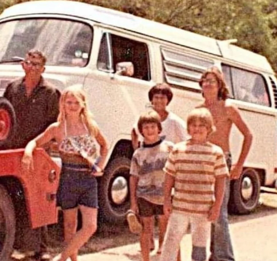 A vintage photo of six people, including children and adults, standing in front of a white van on a sunny day. Trees are visible in the background. The group looks relaxed and casual, dressed in summer clothes.
