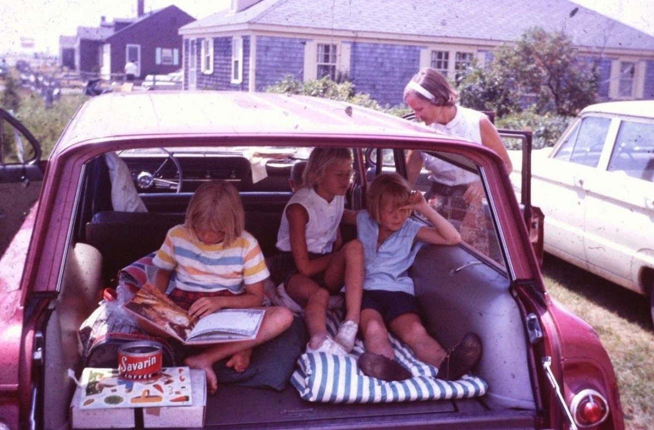 Three children sit in the open trunk of a red station wagon, reading and relaxing, while an adult woman stands nearby. The scene is set outside a house, with another car parked beside them.