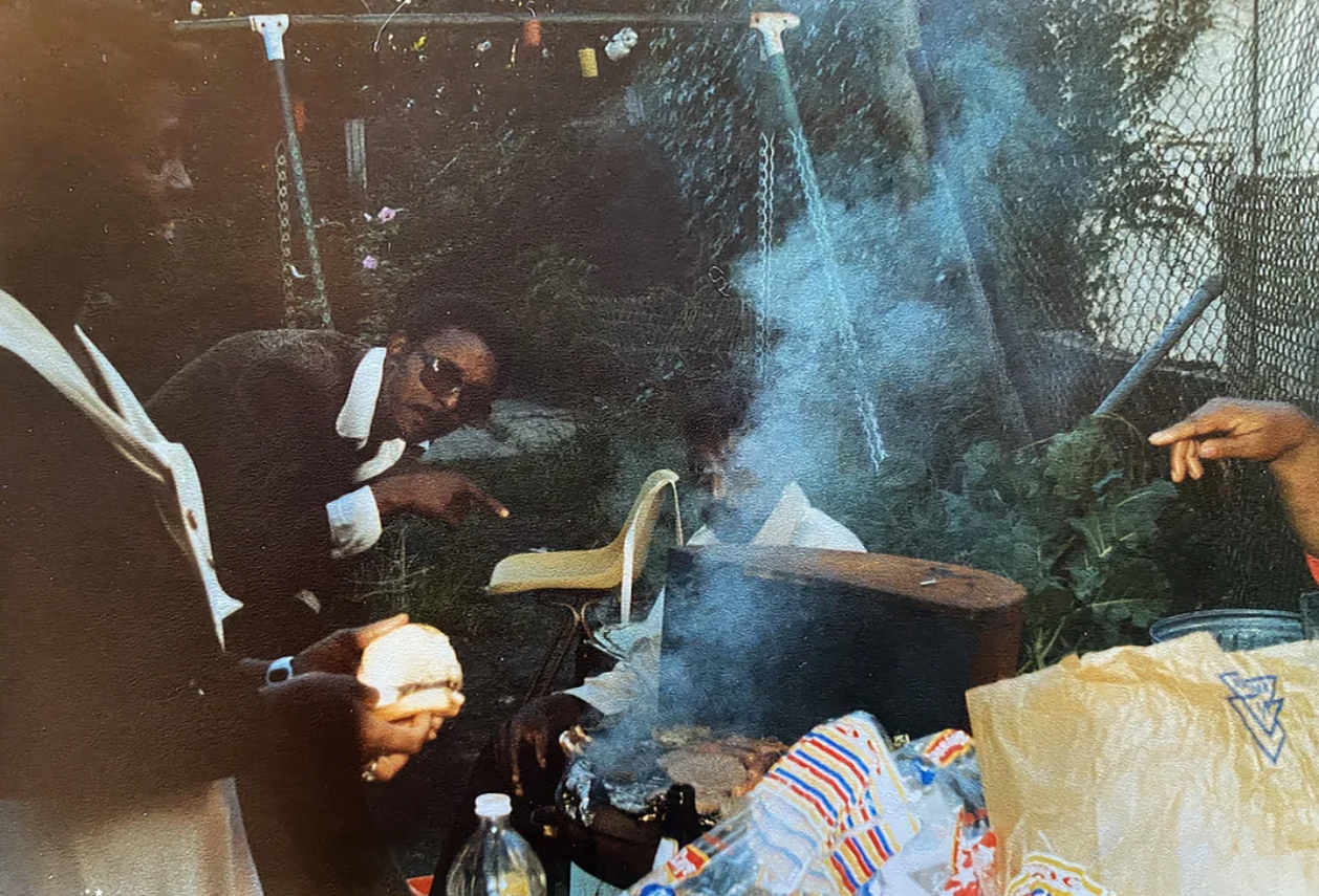 Three people gather around a smoky grill outdoors, with one person pointing and another holding food. A swing set, foliage, and a shopping bag are visible in the background. The scene appears candid and lively.