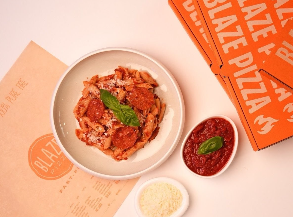 A bowl of pasta with pepperoni and basil, a small dish of tomato sauce with basil, grated cheese, Blaze Pizza menu, and stacked Blaze Pizza boxes—showcasing one of the standout newer pizza chains on a light background.