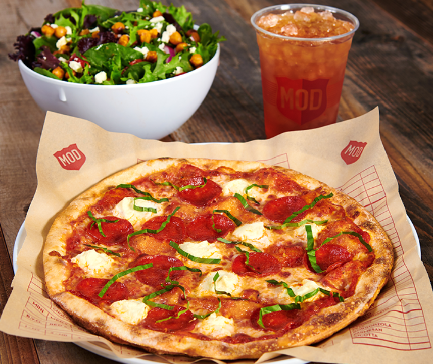 A pepperoni pizza with basil and dollops of cheese on a MOD-branded plate—a standout among newer pizza chains—served with a mixed green salad topped with chickpeas and a cup of iced tea on a wooden table.
