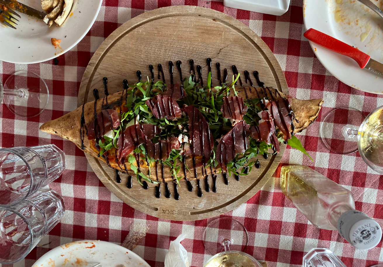 A baked bread dish reminiscent of creations from newer pizza chains is topped with greens, sliced cured meat, and balsamic glaze, sitting on a wooden board atop a red-and-white checkered tablecloth amid plates and glasses.