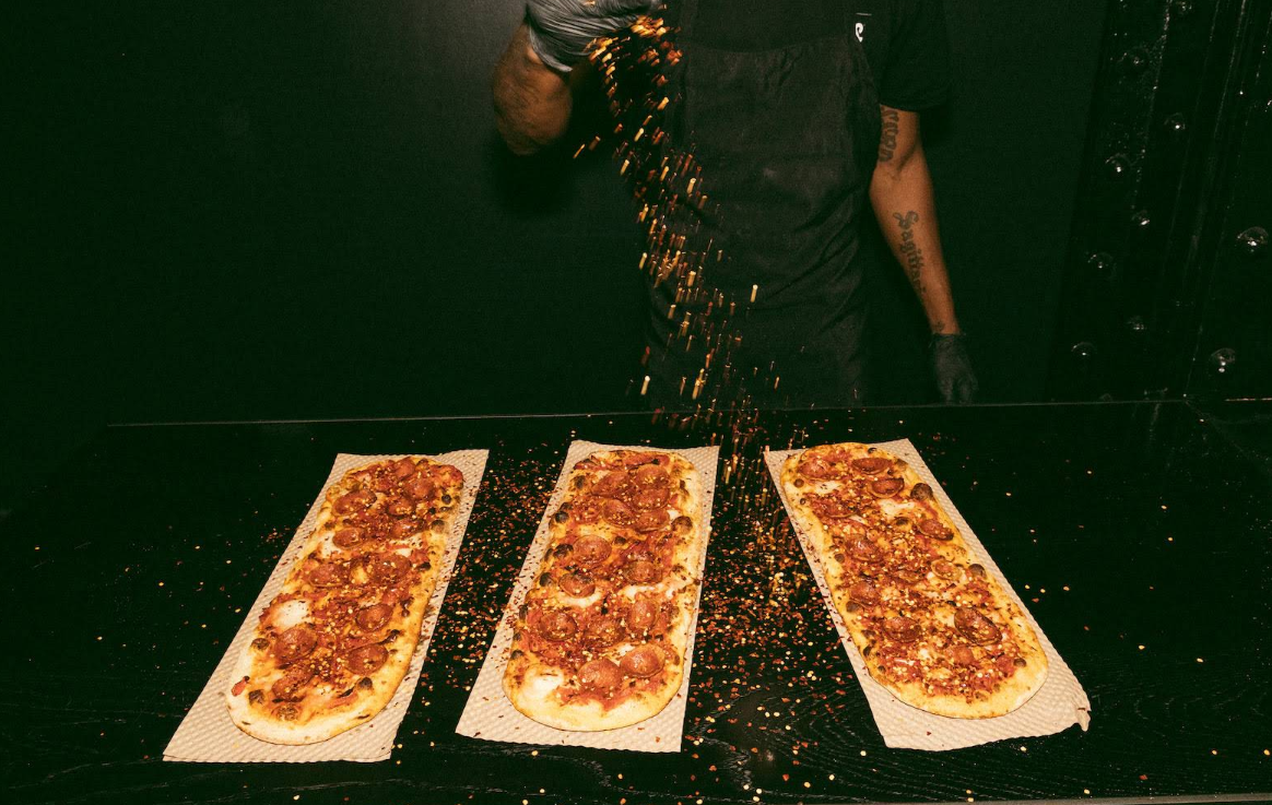A person wearing black gloves sprinkles seasoning over three long, rectangular pizzas topped with pepperoni from one of the newer pizza chains, set on a dark surface against a dark background.
