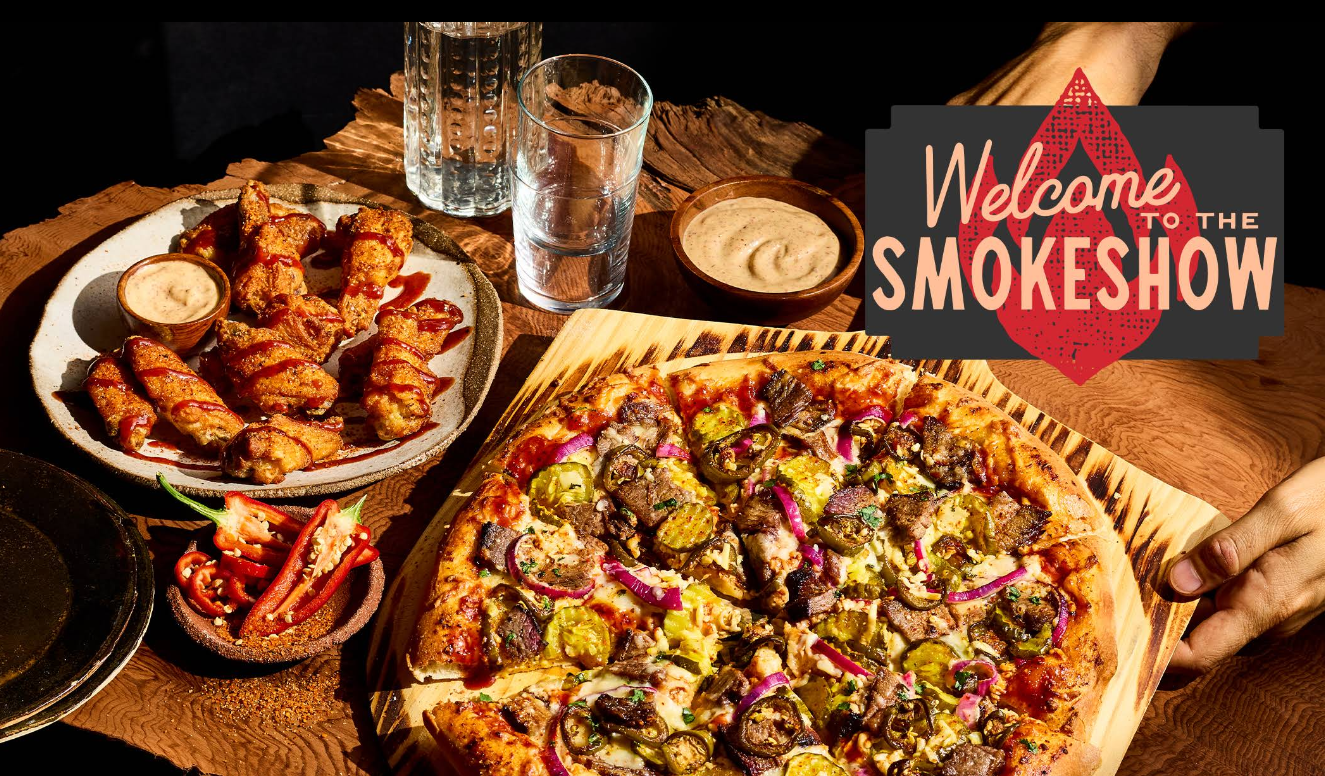 A wood-fired pizza from one of the newer pizza chains, topped with meat, pickles, and red onions, sits on a wooden table next to chicken wings, dipping sauce, cut vegetables, and glasses of water. A sign reads “Welcome to the Smokeshow.”