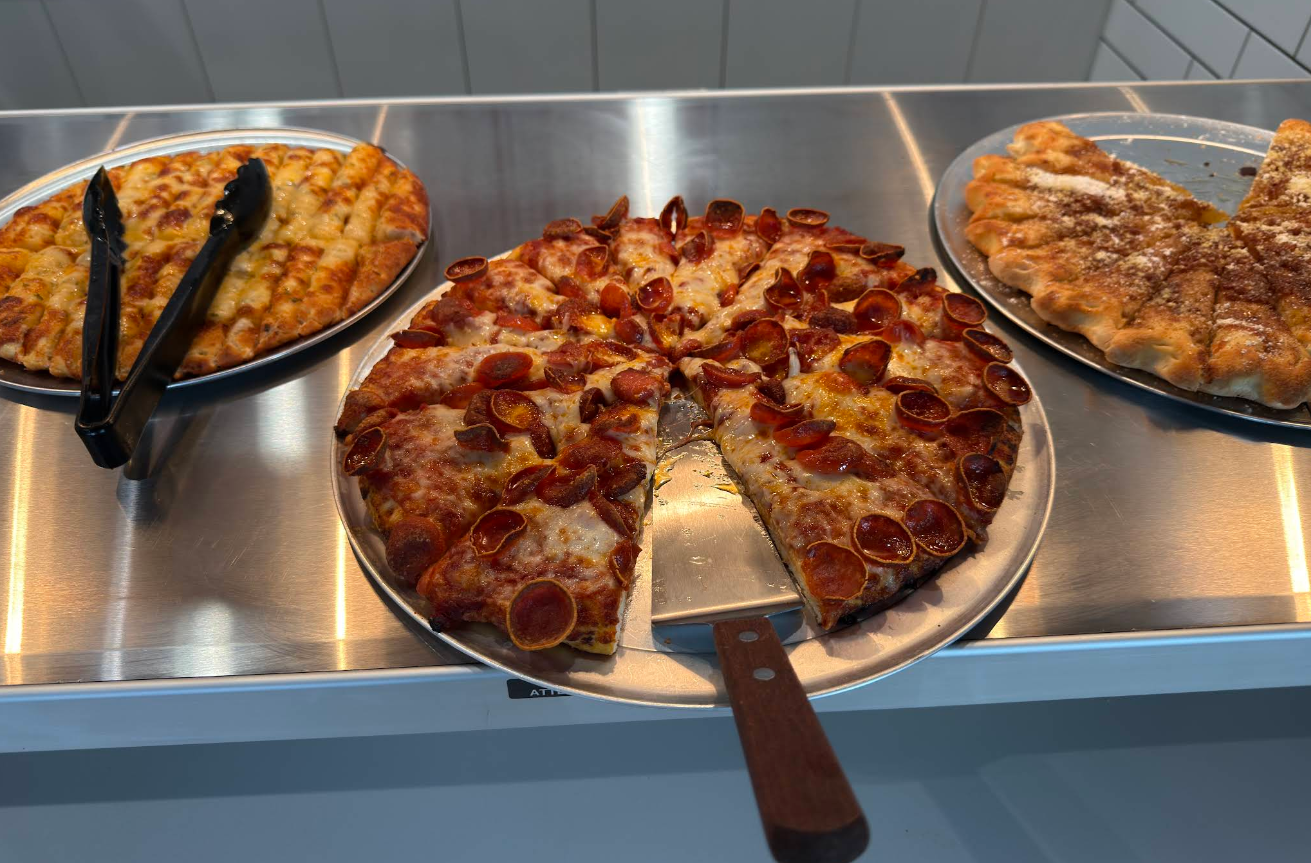 Three metal trays display different types of pizza and breadsticks on a counter. The center tray holds pepperoni pizza slices, while the side trays, inspired by newer pizza chains, feature cheesy breadsticks and cinnamon-sugar breadsticks.