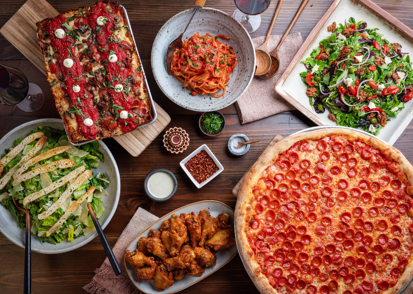 A table set with a variety of Italian-American foods from newer pizza chains, including pepperoni pizza, lasagna, spaghetti, chicken wings, Caesar salad, mixed green salad, and glasses of red wine.