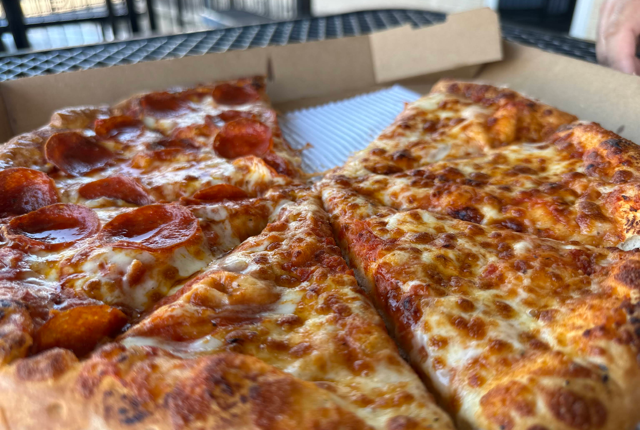 A close-up of a pizza in an open box from one of the newer pizza chains, showing half with pepperoni slices and the other half with just melted cheese, on a metal table outdoors.