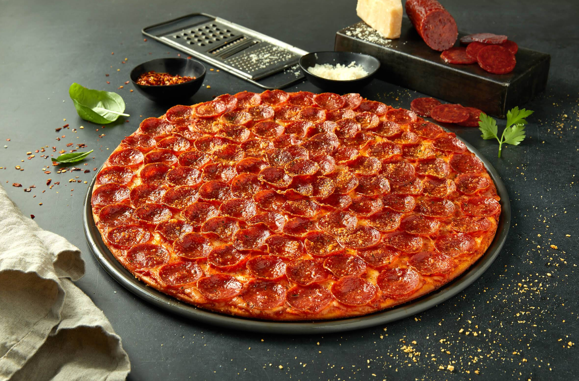 A large pepperoni pizza from one of the newer pizza chains rests on a dark surface, surrounded by a grater, grated cheese, a block of cheese, sliced sausage, herbs, crushed red pepper, and a beige napkin.