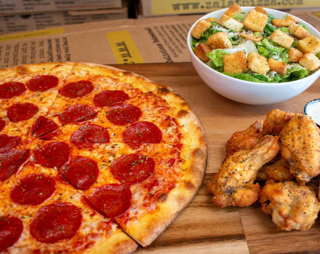 A pepperoni pizza from one of the newer pizza chains, a bowl of Caesar salad with croutons, and several pieces of seasoned chicken wings with ranch dip are arranged on a wooden surface.