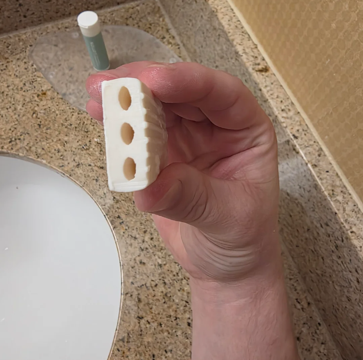 A hand holds a white rectangular object with three evenly spaced holes, over a bathroom countertop near a sink. A small bottle and the drain are visible in the background.