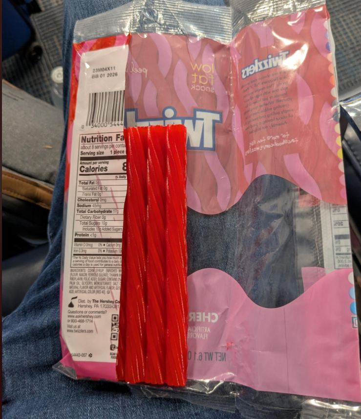 A package of Twizzlers candy rests on someone's lap, with a few red licorice twists visible. The package is partially open, showing the nutrition facts label and the cherry flavor. Blue jeans are visible underneath.