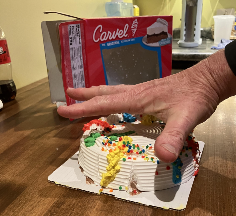 A hand is pressing down on a partially smashed Carvel ice cream cake with colorful icing and sprinkles, next to its open box on a wooden countertop. A bottle of soda and kitchen items are in the background.