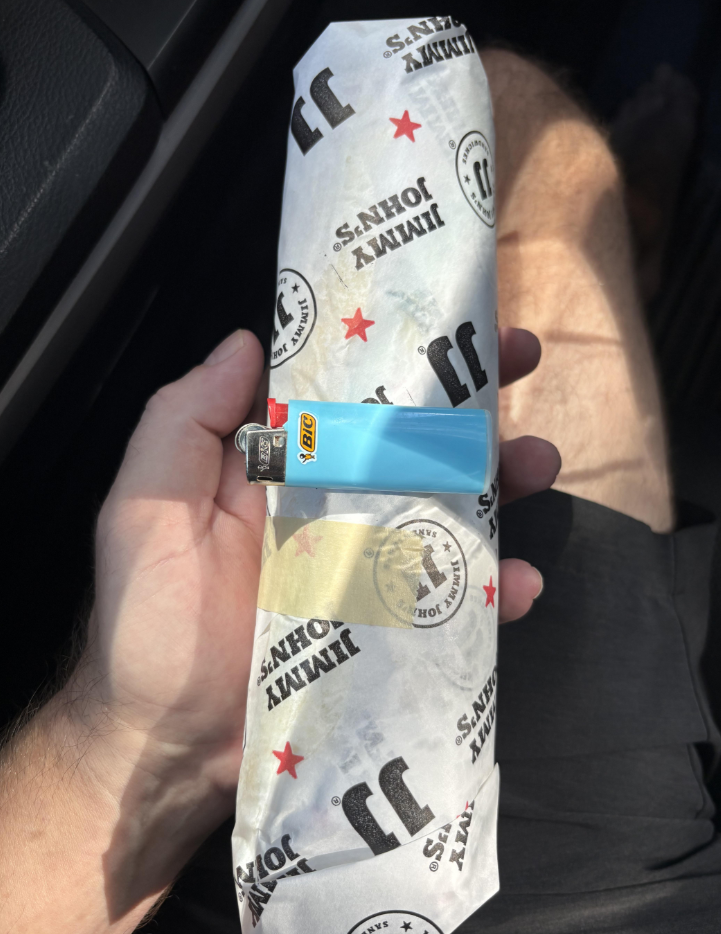 A hand holds a wrapped Jimmy John's sandwich with a blue Bic lighter resting on top, highlighting the large size of the sandwich. The person is wearing black shorts and sitting in a car.