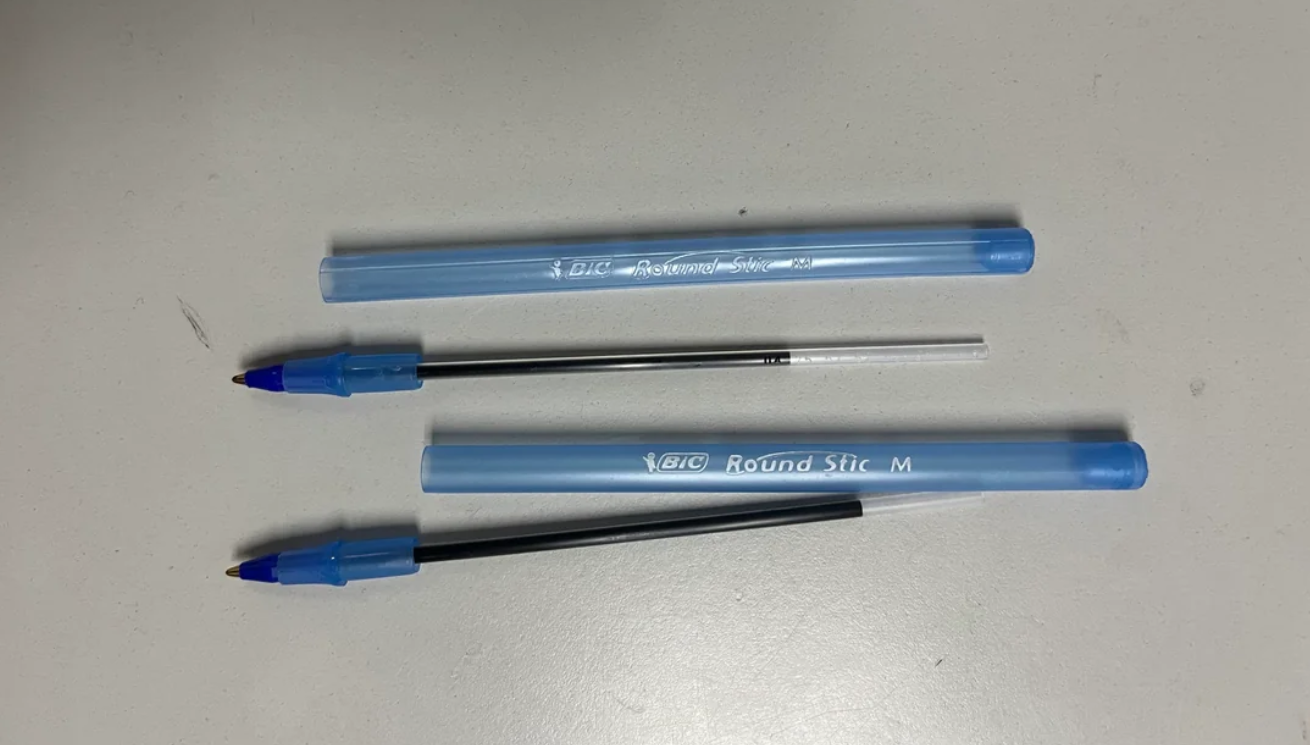 Two blue BIC Round Stic pens are lying open on a white surface. One pen has a black ink refill, and the other has a clear, empty ink refill placed beside its barrel and cap.