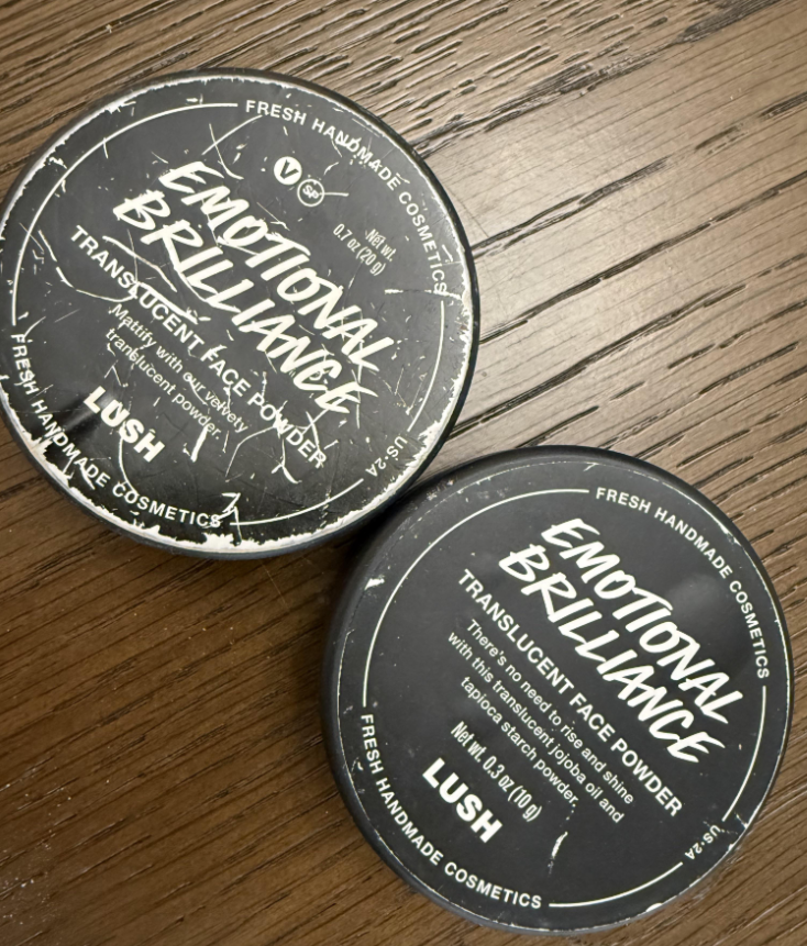 Two round black containers of Lush Emotional Brilliance Translucent Face Powder are placed on a wooden surface. The containers have white text and appear slightly worn.