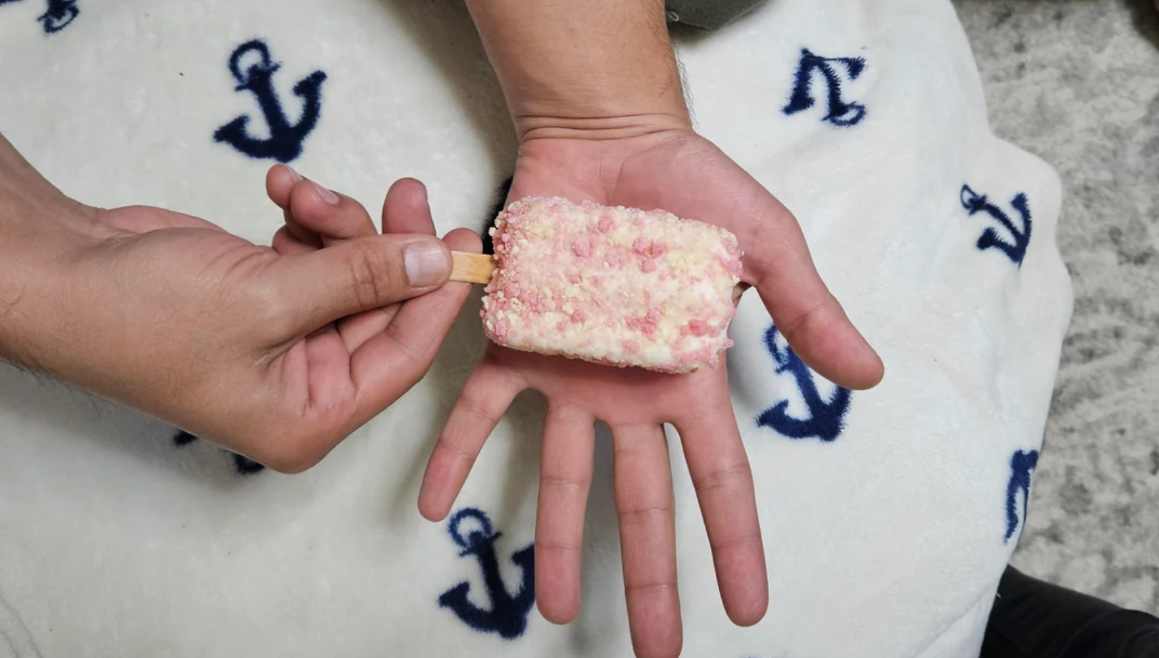 A person holding a rectangular pink and white ice cream bar on a stick over their open palm, with a white blanket featuring blue anchor patterns in the background.