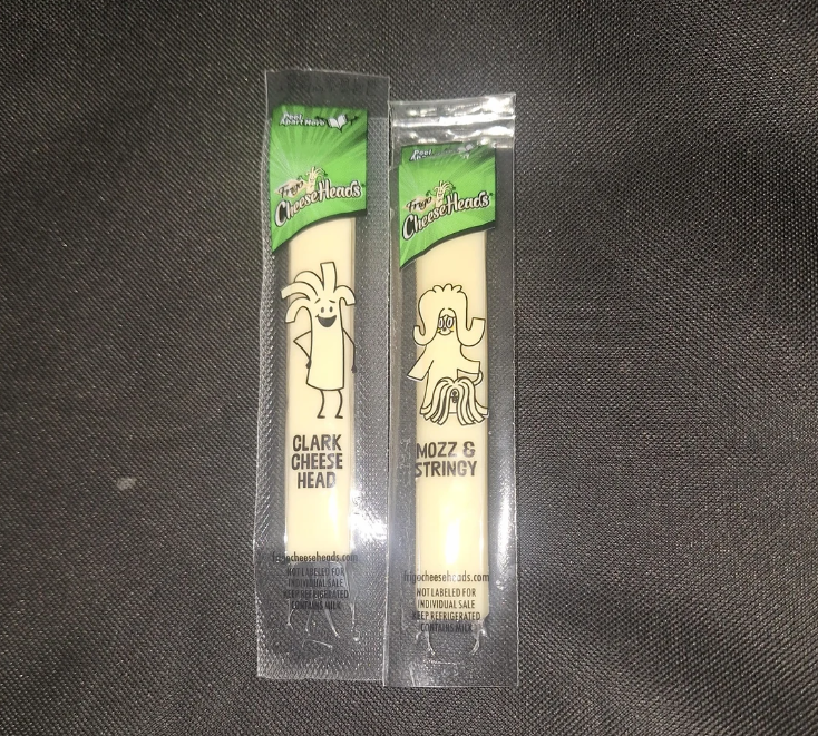 Two individually wrapped string cheese sticks on a dark fabric background. Each wrapper features a cartoon cheese character: one labeled "Clark Cheese Head" and the other "Mozz & Stringy.