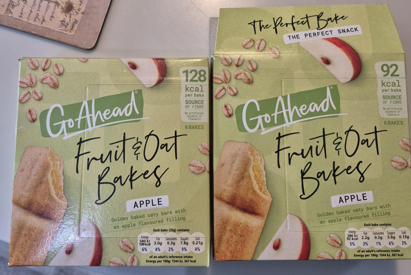 Two green boxes of Go Ahead Fruit & Oat Bakes, Apple flavor, are side by side on a counter. The left box shows "128 kcal" per bake; the right shows "92 kcal". Both feature images of the oat bars and oats.