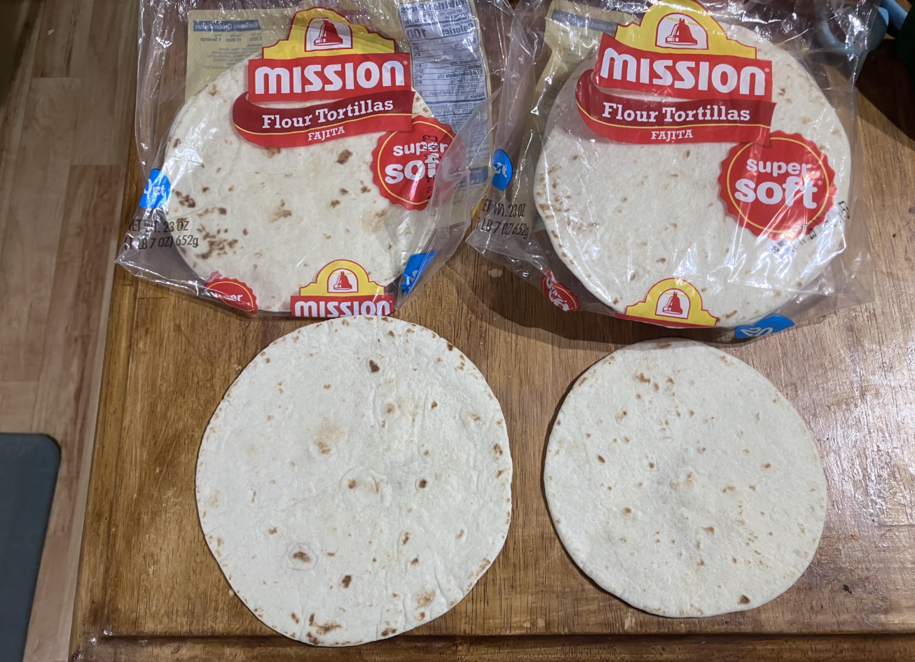 Two packs of Mission Flour Tortillas are on a wooden surface, each with an opened tortilla placed in front, showing the round, soft tortillas outside their clear packaging.