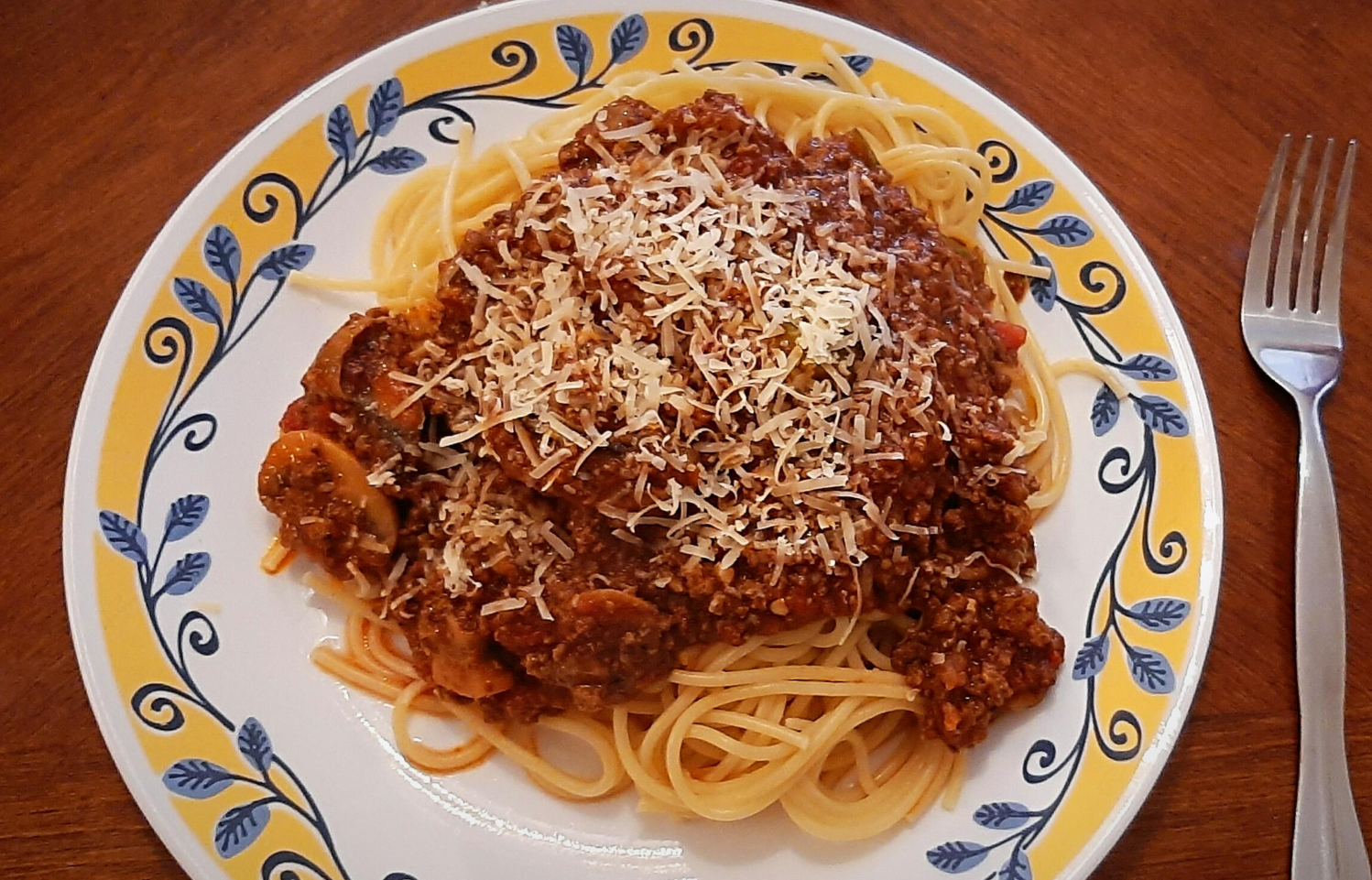 A plate of spaghetti topped with meat sauce and grated cheese, a staple of classic dinners, sits on a decorative plate with a yellow and blue pattern. A fork rests to the right on a wooden table.