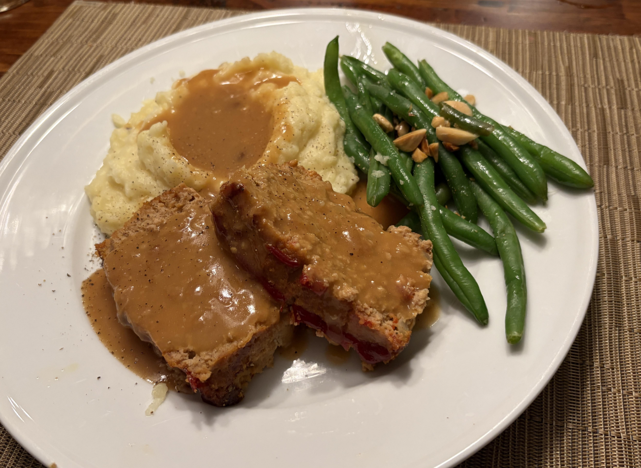 A white plate with two slices of meatloaf topped with brown gravy, mashed potatoes with gravy, and green beans garnished with slivered almonds sits on a woven placemat—a comforting favorite among classic dinners.