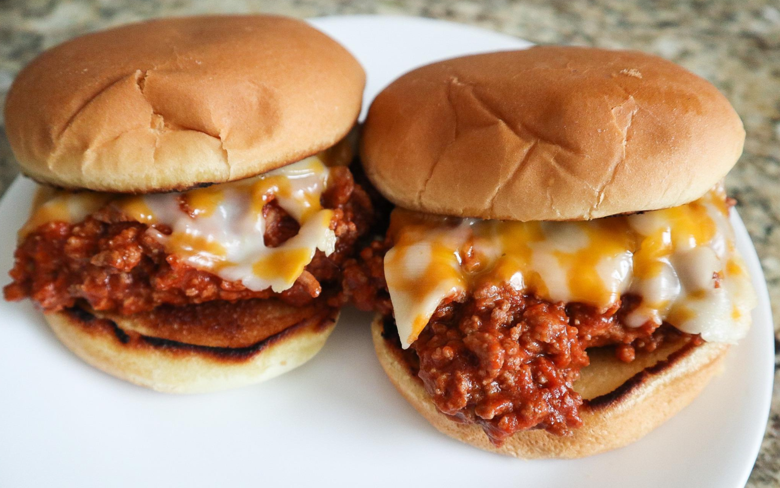 Two sloppy joe sandwiches with melted cheese on top, served on soft hamburger buns and placed on a white plate, make for one of those classic dinners perfect for any kitchen countertop scene.