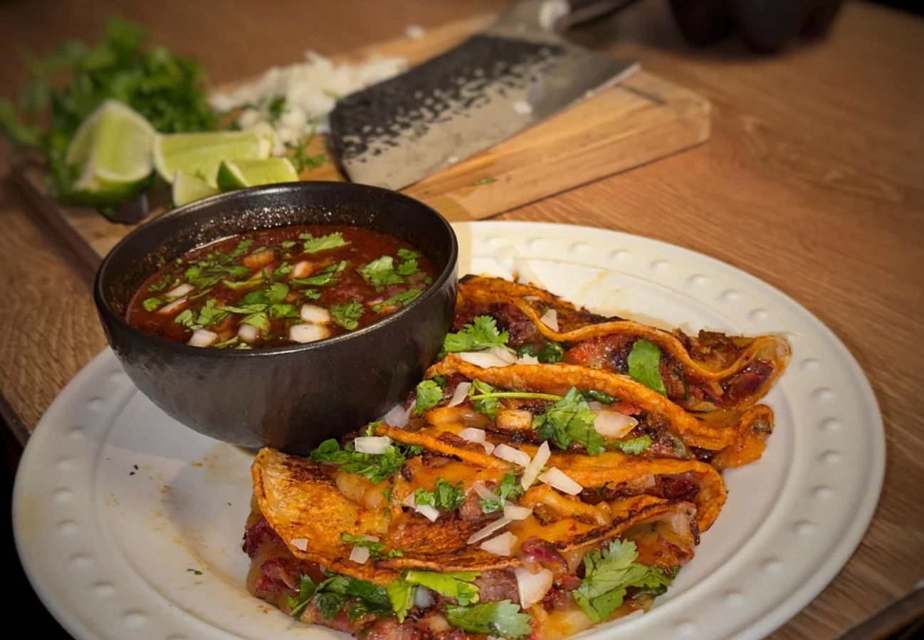 Three birria tacos topped with cilantro and onions are served on a white plate beside a bowl of consommé. In the background, chopped onions, lime wedges, and a cutting board evoke the comfort of classic dinners.