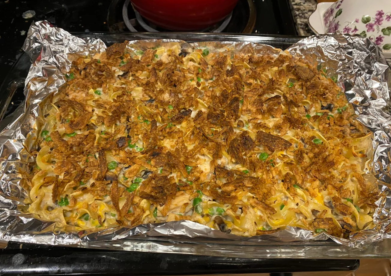 A casserole dish lined with foil, filled with a baked noodle casserole—one of those classic dinners—topped with crispy fried onions, peas, and bits of vegetables, sitting on a stovetop.